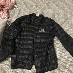 Svart pufferjacka från Emporio Armani - Säljer en snygg svart pufferjacka från Emporio Armani EA7. Jackan har en stilren design med dragkedja och är perfekt för kyligare dagar. Den är i bra skick och har en logga på bröstet samt på dragkedjan. Perfekt för både vardag och fest!