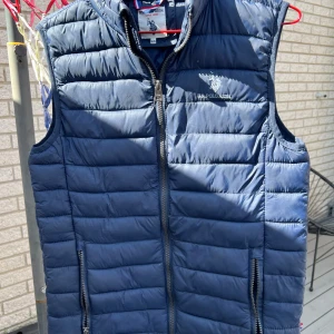 Blå quiltad dunväst från U.S. Polo Assn. - Säljer en snygg blå quiltad dunväst från U.S. Polo Assn. Västen har en dragkedja framtill och är perfekt för höst- och vårsäsongen. Den är i bra skick och har en stilren design med märkets logga på bröstet. Perfekt för lager-på-lager-stil!