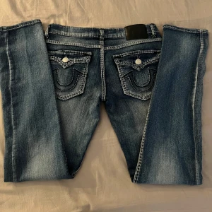 Blå jeans med detaljerade bakfickor - Säljer ett par snygga lågmidjade True Religion jeans med coola detaljer på bakfickorna. De har en klassisk femficksdesign och en snygg tvätt som ger dem en vintage look. Endast använda en gång, finns inga defekter 💞säljer pga att de är lite stora. Midjemåttet är 37 cm och innebärslängden 82 cm