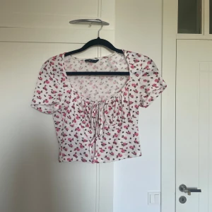 Blommig croppad topp med knyt - Söt och somrig blommig topp med korta ärmar och knyt framtill. Den är croppad och har ett fint mönster med små rosa blommor på en vit bakgrund. 