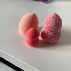 Tre olika beautyblenders i rosa nyanser - Säljer tre olika beautyblenders i olika rosa nyanser. Den första är en klassisk äggformad beautyblender i ljusrosa, perfekt för att applicera foundation jämnt. Den andra är en mindre, rundad beautyblender i en djupare rosa färg, idealisk för concealer eller highlighter. Den tredje har en unik form med två rundade sektioner, vilket gör den mångsidig för olika delar av ansiktet. Alla är mjuka och lätta att använda för en felfri sminkbas.
