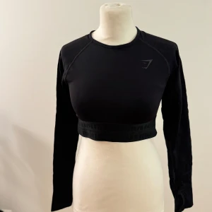 Svart croppad tröja från Gymshark - Säljer en snygg och stilren svart croppad tröja från Gymshark. Den är perfekt för träning eller en avslappnad dag. Tröjan har långa ärmar och en tight passform som framhäver figuren. Materialet är mjukt och stretchigt för bästa komfort. Perfekt för dig som vill ha en sportig look!