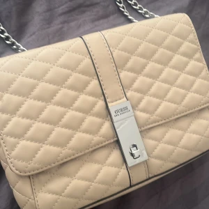 Beige quiltad axelväska från Guess - Säljer en superfin beige axelväska från Guess. Den har en snygg kedja som axelrem. 