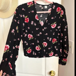 Svart blommig blus - Säljer en superfin svart blus med röda blommor och små rosa detaljer. Blusen har långa ärmar och en v-ringad hals. Perfekt för både vardag och fest! Den är långärmad och jätrebekväm. Köpt för 299kr säljer för endast 99kr