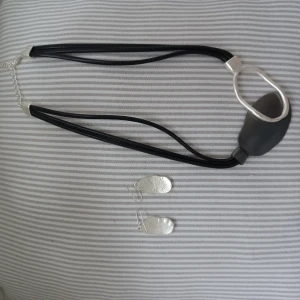 Svart halsband och silverörhängen - Elegant halsband med svart rem och en stor silverfärgad detalj. Matchande örhängen i silver med unikt mönster. Perfekt för att ge en stilren touch till din outfit.
