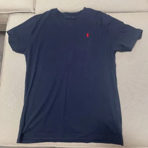Mörkblå t-shirt från Ralph Lauren - Säljer en klassisk mörkblå t-shirt från Ralph Lauren med det ikoniska röda broderade logotypen på bröstet. Perfekt för en stilren och avslappnad look. T-shirten är kortärmad och tillverkad i mjuk bomull. Storlek står XL i barn storlek men motsvarar S/M