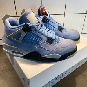 Jordan 4 UNC - Snygga Nike Air Jordans i ljusblå mocka med svarta och grå detaljer. Skorna har en klassisk design med snörning och en synlig Air-enhet i sulan för extra komfort. Perfekta för dig som vill ha en stilren och sportig look.