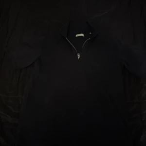 Svart pikétröja från Jack & Jones - Säljer en stilren svart pikétröja från Jack & Jones med dragkedja i halsen. Perfekt för en avslappnad men ändå snygg look. Tröjan har korta ärmar och är gjord i ett bekvämt material.