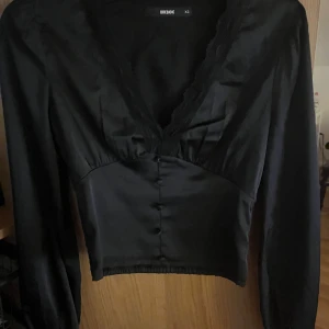 Svart blus från Bik Bok - Snygg svart blus från Bik Bok i satin med v-ringning och långa ärmar. Blusen har knappar framtill och spetsdetaljer vid halsen. Perfekt för en fest eller bara en middag. Säljer pågrund av att den är för liten nu då den hängt i garderoben efter en användning. Priset går att förhandla💞