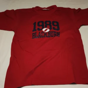 Röd t-shirt med tryck - Säljer en röd t-shirt med trycket '1989 Blackburn' i svart. T-shirten har korta ärmar och en rund halsringning. Perfekt för en avslappnad stil.