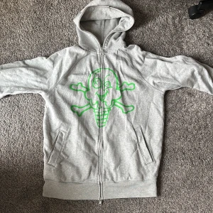Grå hoodie från billionaire boys club  - Säljer en grå hoodie från billionaire boys club med ett coolt grönt tryck av en glass och korsade benknotor på framsidan. Hoodien har dragkedja och två fickor framtill. Perfekt för en avslappnad stil. Nästan aldrig använd 