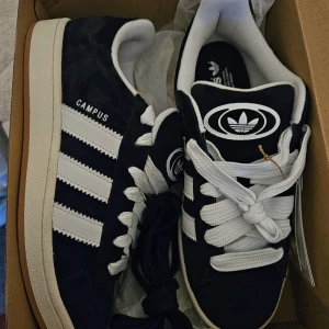 Svarta Adidas Campus sneakers - Snygga svarta Adidas Campus sneakers med vita ränder och snörning. Skorna har en klassisk design med gummisula och är perfekta för en avslappnad stil. De har en bekväm passform och är ett måste för alla sneaker-älskare.
