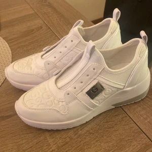 Vita sneakers med broderad detalj - Snygga vita sneakers med en unik broderad detalj på ovansidan. Skorna har en diskret metallisk logotyp på sidan och en bekväm passform. Perfekta för en stilren look. Köpta i USA använda endast 1 gång