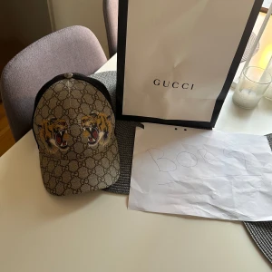 Tiger keps från Gucci - Säljer en stilren keps från Gucci med deras ikoniska mönster och två broderade tigrar framtill. Kepsen har en justerbar rem baktill. Enda som är fel är att tvättlappen lossnat. Finns med alla tillbehör.