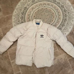 Vit pufferjacka från Adidas - Säljer en snygg vit pufferjacka från Adidas med dragkedja och det klassiska logotyptrycket på bröstet. Perfekt för kyliga dagar!