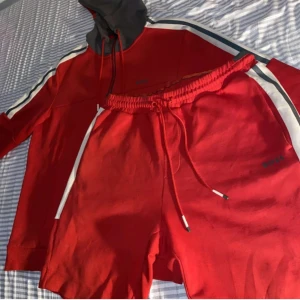 Röd Hoodie matchande shorts set ifrån BOSS.  - Snygga röda shorts och Kofta från Boss med vita detaljer längs sidorna. De har en elastisk midja med snörning för justerbar passform. Perfekta för en sportig look eller avslappnad stil.