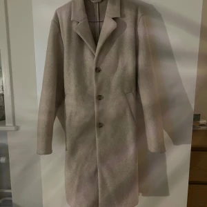 Beige kappa - Elegant beige kappa med klassisk design och knappar framtill från H&M. Den har långa ärmar och en stilren krage, perfekt för kyligare dagar. Passar bra till både vardag och mer uppklädda tillfällen. Den är knappt använd