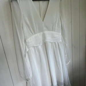 Vit klänning med knytband - Elegant vit klänning med v-ringning och långa ärmar. Den har ett knytband i midjan som ger en fin siluett. Perfekt för en stilren look.