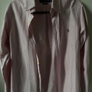 Randig skjorta från Ralph Lauren - Klassisk långärmad skjorta från Ralph Lauren med smala ränder. Finns i både rosa och blå variant. Skjortan har knappar framtill och en broderad logotyp på bröstet. Perfekt för en stilren look.
