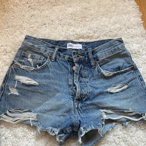 Blå jeansshorts från Zara - Snygga blå jeansshorts från Zara med slitna detaljer och fransig kant. De har en knappgylf och klassisk femficksdesign. Midwaist men kan också bäras highwaist eller low waist. Perfekta för en avslappnad stil.