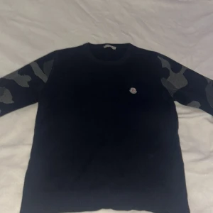 Svart tröja med camoärmar från Moncler - Snygg svart tröja från Moncler med coola camo-detaljer på ärmarna. Perfekt för en stilren look med en twist. Tröjan har en rund hals och Moncler-logga på bröstet.