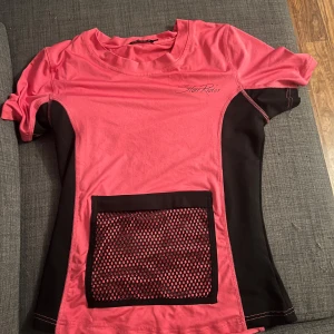 Rosa och svart ridtröja från Star Rider - Snygg rosa och svart ridtröja från Star Rider med korta ärmar och ett nätficka framtill med dragkedja perfekt för att ha telefonen i. 