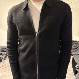 Svart tröja med dragkedja från Samsøe Samsøe - Snygg svart tröja från Samsøe Samsøe med dragkedja framtill. Tröjan har en klassisk krage och en bröstficka. Perfekt för en stilren look. Nypris 1400kr