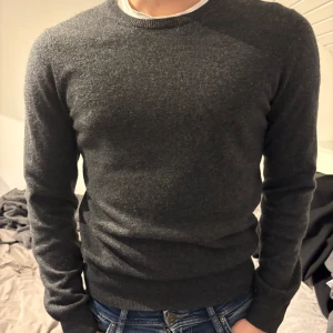 Mörkgrå tröja från Uniqlo - Säljer en stilren mörkgrå tunnare tröja från Uniqlo. Tröjan har en klassisk rund hals och långa ärmar, perfekt för kyligare dagar. Den är mjuk och bekväm, vilket gör den till ett måste i garderoben. Passar både till jeans och mer uppklädda tillfällen.