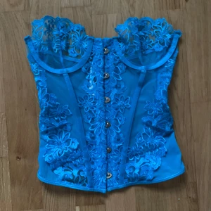 Blå spetskorsett från PrettyLittleThing - Säljer en snygg blå korsett från PrettyLittleThing med vackra spetsdetaljer och knappar framtill. Den har en transparent design med blommönster och justerbara axelband. Perfekt för att ge en extra touch till din outfit!