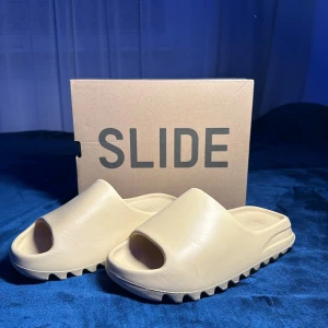 Beige Adidas Yeezy Slide - Säljer ett par beige Adidas Yeezy Slide. Dessa sandaler har en minimalistisk design med en bekväm passform och räfflad sula för extra grepp. Perfekta för en avslappnad stil. Priset går att sänka 