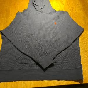 Marinblå hoodie från Ralph Lauren - Säljer en snygg marinblå hoodie från Ralph Lauren med det klassiska logotypen i orange på bröstet. Tröjan har en skön huva och en praktisk magficka. Perfekt för en avslappnad stil.