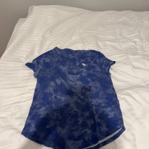 Blå t-shirt från Abercrombie Kids - Säljer en blå t-shirt från Abercrombie Kids med ett mönster som påminner om moln. T-shirten har korta ärmar och en liten logga på bröstet. Perfekt för en avslappnad stil.