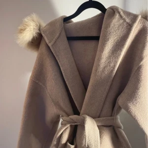 Beige kappa med pälsdetalj - Elegant beige kappa med en lyxig pälsdetalj runt huvan. Kappan har en knytning i midjan för en smickrande passform. Perfekt för kyliga dagar när du vill hålla stilen.