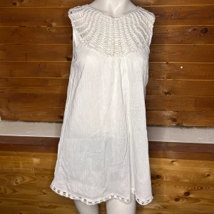 Vit ärmlös klänning med virkad detalj - Säljer en vit ärmlös topp med en vacker virkad detalj runt halsen. Toppen har en luftig passform och är perfekt för varma dagar. Den är tillverkad i ett lätt och bekvämt material.