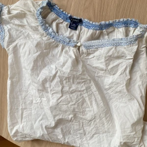 Vit blus från Gant med blå detaljer - Säljer en söt vit blus från Gant med blå spetsdetaljer vid hals och ärmar. Blusen har korta ärmar och en avslappnad passform. Perfekt för en somrig look! 🌼