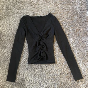 Svart långärmad topp från BikBok - Snygg svart långärmad topp från BikBok i storlek XS. Toppen har en v-ringad design med knytning framtill som ger en elegant touch. Perfekt för både vardag och fest!