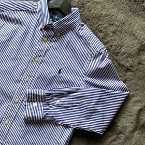 Randig skjorta från Ralph Lauren - Säljer en klassisk blå- och vitrandig skjorta från Ralph Lauren i storlek M. Skjortan har långa ärmar och knappar framtill, perfekt för både vardag och fest. Den är i mycket bra skick och gjord av bomull, vilket gör den bekväm och stilren. Passar perfekt till både jeans och chinos!