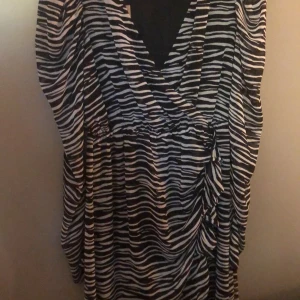 Zebramönstrad omlottklänning - Snygg zebramönstrad omlottklänning i svart och vitt. Perfekt för en utekväll eller fest! Klänningen har en v-ringad design och är i ett mjukt och bekvämt material. Den har en smickrande passform med knytning i midjan. Ny med prislappen kvar! Aldrig använd. 