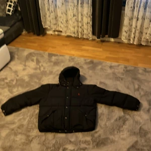 Svart dunjacka från Polo Ralph Lauren - Säljer en snygg svart dunjacka från Polo Ralph Lauren i storlek L. Jackan har en broderad röd logga på bröstet och är perfekt för kalla vinterdagar. Den är långärmad och har knappar samt dragkedja för extra skydd mot kylan. Passar både till vardags och festliga tillfällen.Jackan är använd nått år skriv gärna för mer bilder på jackan.Pris kan diskuteras vid snabb affär