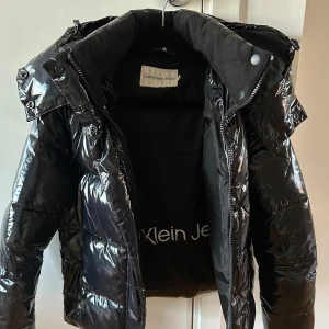 Svart pufferjacka från Calvin Klein Jeans - Säljer en snygg svart pufferjacka från Calvin Klein Jeans i storlek XS köpt förra vintern . Jackan är i mycket bra skick använd 2-3 gånger och perfekt för vintern. Den har en glansig yta med dragkedja och knappar, samt en mysig huva. Perfekt för att hålla sig varm och stilren under kalla dagar!       Pris kan diskuteras vid snabb affär kan mötas i Stockholm annars postas den. Köpt för 2200kr