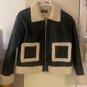 Svart jacka med fuskpälsdetaljer - Säljer en snygg svart jacka från Ginatrico med mysiga fuskpälsdetaljer i vitt. Jackan har en dragkedja framtill och är perfekt för höst och vinter. Den har en normal passform och långa ärmar. Superfin att ha både till vardags och fest!