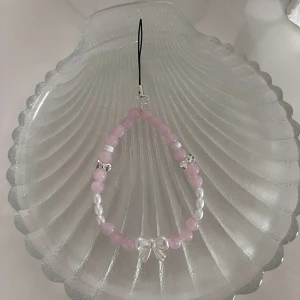 Rosa och vitt mobilsmycke med pärlor - Snyggt mobilsmycke med rosa och vita pärlor, perfekt för att ge en söt touch till din outfit. Accesoaren har en liten rosett i mitten. Perfekt för både vardag och festliga tillfällen!