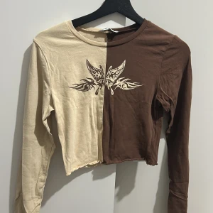 Beige och brun croppad tröja från FB Sister - Säljer en cool croppad tröja från FB Sister i beige och brun. Den har ett unikt fjärilstryck på framsidan och är långärmad. Perfekt för en avslappnad stil med en twist. Passar bra till både jeans och kjol!
