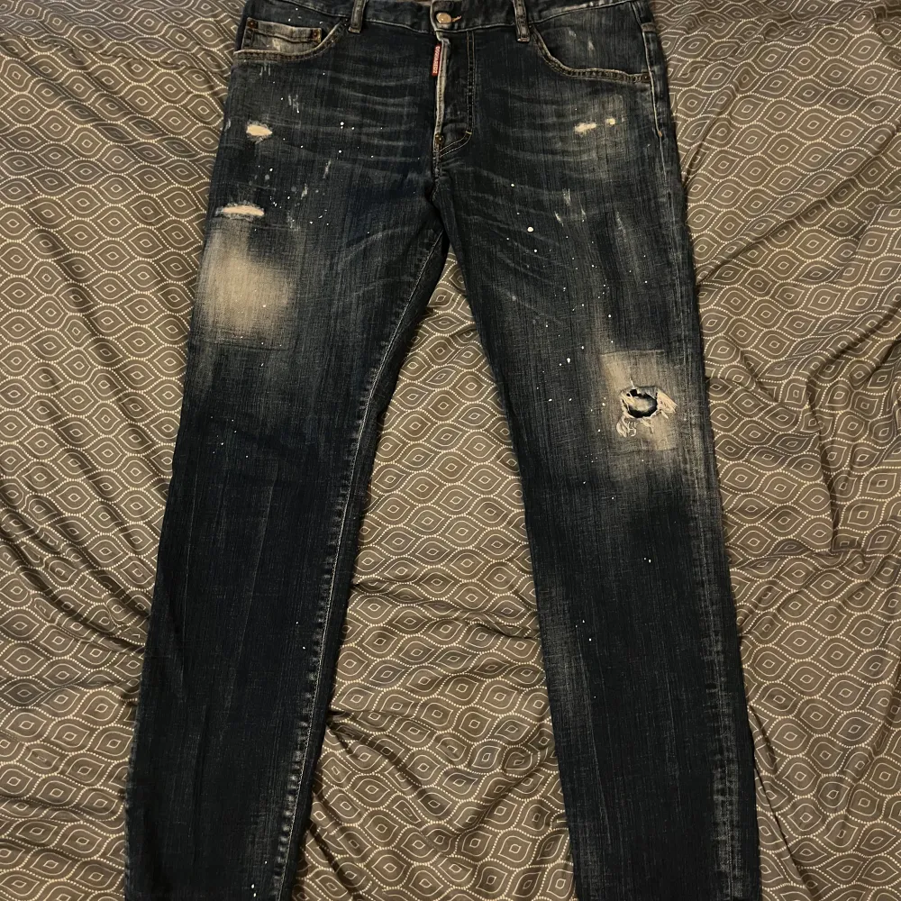 Säljer ett par blå DSQUARED2 jeans i storlek 50. De har en cool distressed look med slitningar och färgstänk. Perfekta för en avslappnad men stilren outfit. Jeansen är i bra skick och har en straight passform. Passar perfekt för höst och vår!. Jeans & Byxor.