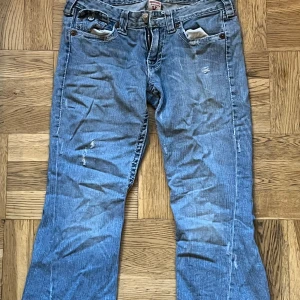 Blå jeans från True Religion - Säljer ett par blå jeans från True Religion i bra skick. De har en snygg slitning och coola detaljer med knappar och broderi på bakfickorna. Perfekta för en avslappnad stil. Passar bra till både vardag och fest! Kontakta gärna innan köp. 