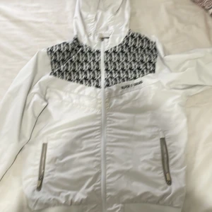 Vit vindjacka från Black Squad - Säljer en snygg vit vindjacka från Black Squad i storlek S. Jackan har en cool svartvit mönstrad detalj på bröstet och en praktisk huva. Den är perfekt för vår och höst med dragkedja och fickor med dragkedjor. Passar både till vardags och lättare aktiviteter.