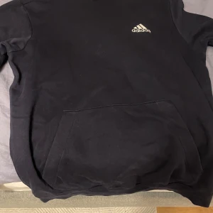 Mörkblå Hoodie från Adidas - Säljer en mörkblå hoodie från Adidas i bra skick. Den har en klassisk design med Adidas-loggan på bröstet och en praktisk magficka. Perfekt för höst och vinter, eller bara för att chilla hemma. Skön och stilren, passar till det mesta! Storlek M 