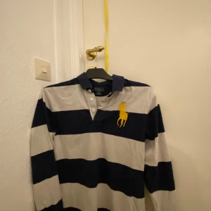 Randig pikétröja från Polo Ralph Lauren - Säljer en klassisk randig pikétröja från Polo Ralph Lauren i blått och vitt. Tröjan har långa ärmar och en stor gul broderad logga på bröstet. Perfekt för en stilren och avslappnad look. Passar både till vardags och lite finare tillfällen. 