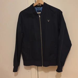 Mörkblå jacka | GANT - Säljer en stilren mörkblå jacka från GANT i mycket bra skick. Jackan har en dragkedja framtill och är långärmad. Perfekt för höst och vår med sin klassiska design och broderade logga på bröstet. Passar både till vardags och lite finare tillfällen.