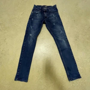 DSquared2 Navy Jeans - Säljer ett par snygga blå jeans från DSquared2 med coola slitningar och detaljer. De har en slim passform och är perfekta för både vardag och fest. Jeansen har en klassisk femficksdesign och en knappgylf. Märkeslogga syns på baksidan. Passar perfekt för dig som vill ha en stilren men ändå edgy look.